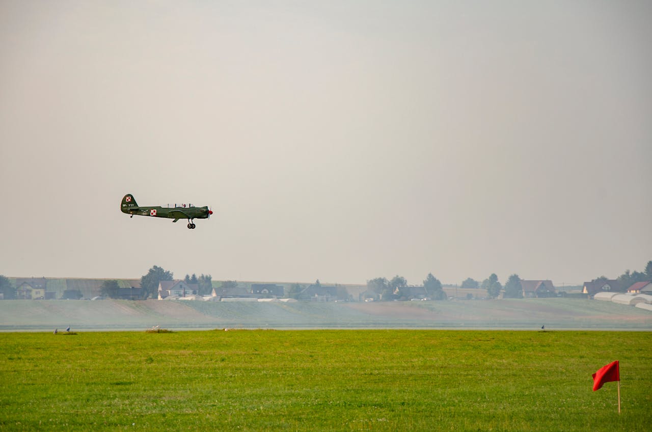 gallery-5 A vintage military aircraft is captured in flight above an airfield with a distant village backdrop.