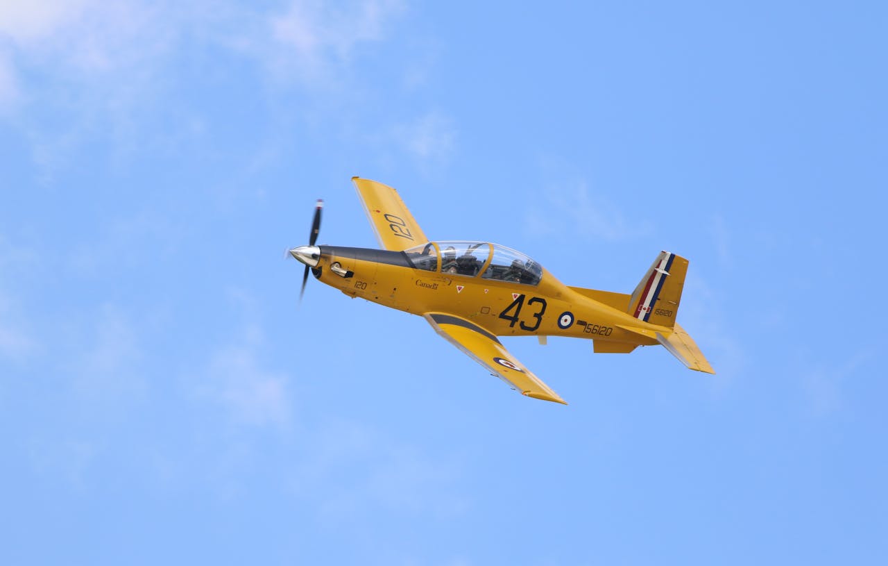 gallery-1 A yellow training aircraft flying high against a vibrant blue sky on a sunny day.
